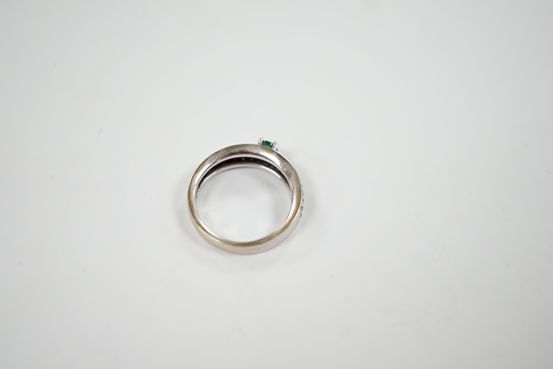 A modern 18ct white gold and single stone oval cut emerald set ring, with pave set diamond shoulders, size L/M, gross weight 4.3 grams.